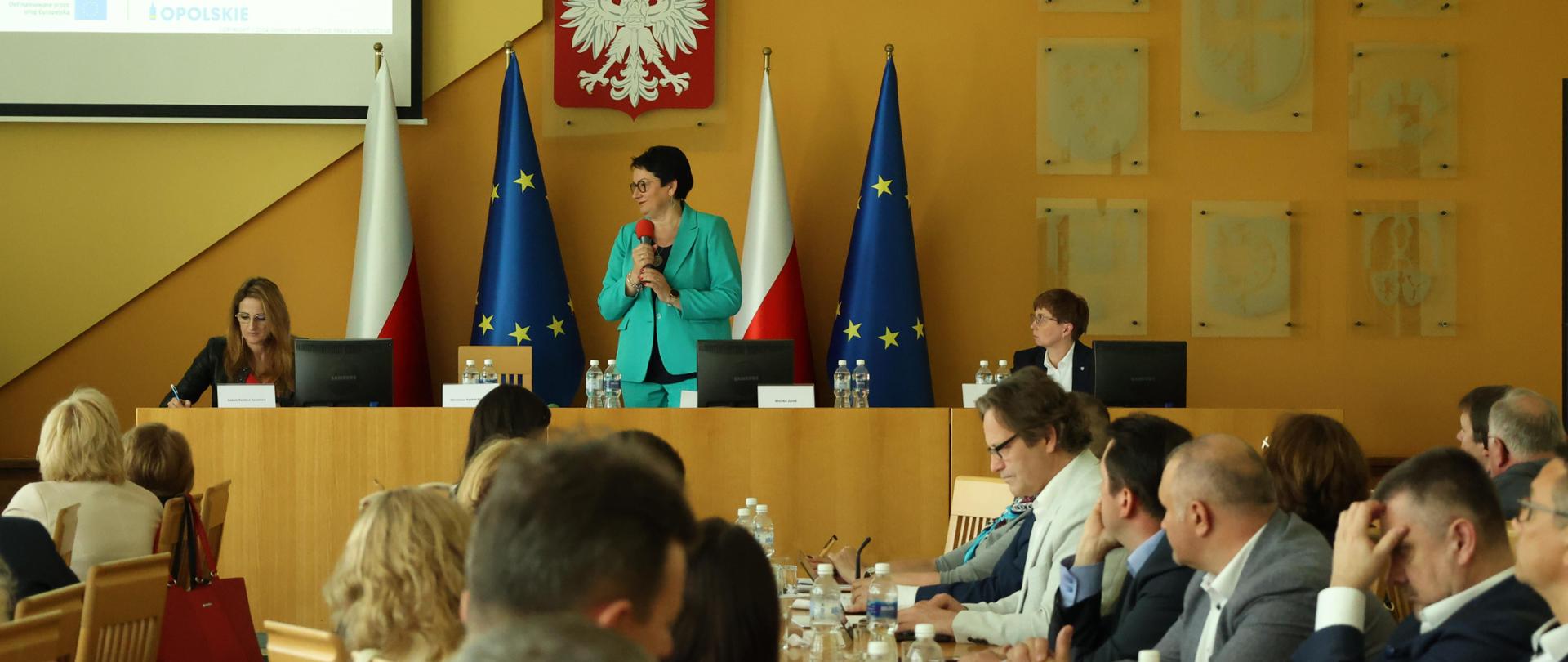 Wojewoda Opolski Monika Jurek podczas narady dotyczącej wojewódzkiego planu transformacji dla województwa opolskiego na lata 2022-2026. Na zdjęciu widać uczestników narady siedzących przy stole oraz Wojewodę, która mówi do mikrofonu. Za Wojewodą znajdują się flagi Polski oraz Unii Europejskiej. 