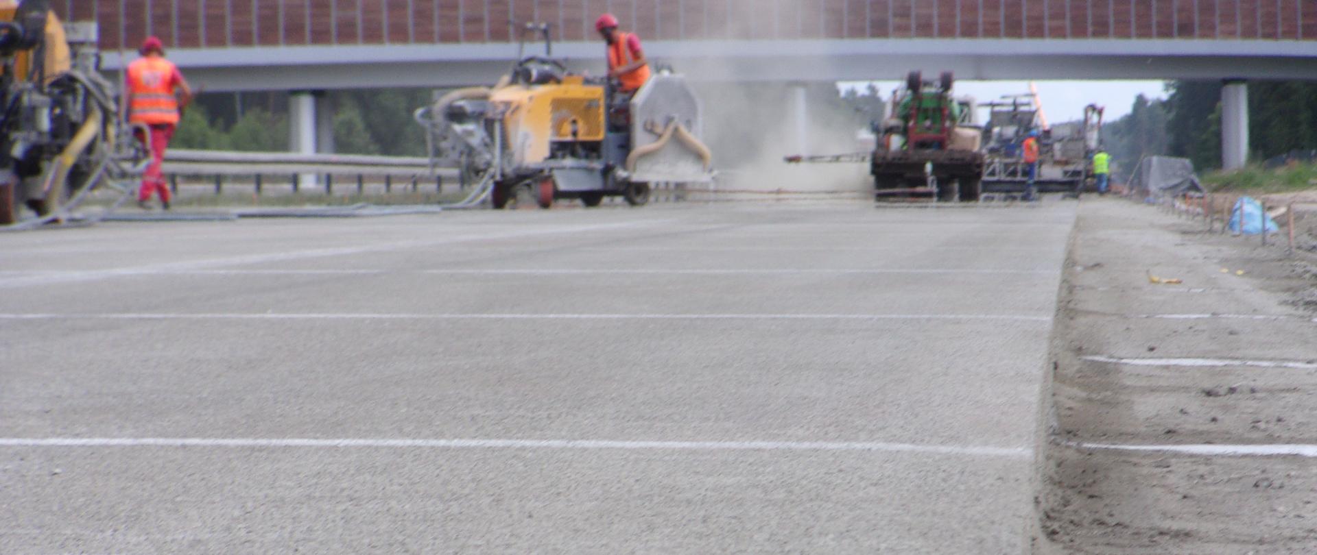 Zdjęcie wykonane z poziomu nawierzchni. Na pierwszym planie widać nawierzchnię betonową z zaznaczonymi miejscami gdzie mają zostać wykonane nacięcia pod szczeliny dylatacyjne. Na dalszym planie ekipa robocza i maszyny drogowe. W oddali nad droga przejście dla zwierząt górne, z panelami drewnianymi. 