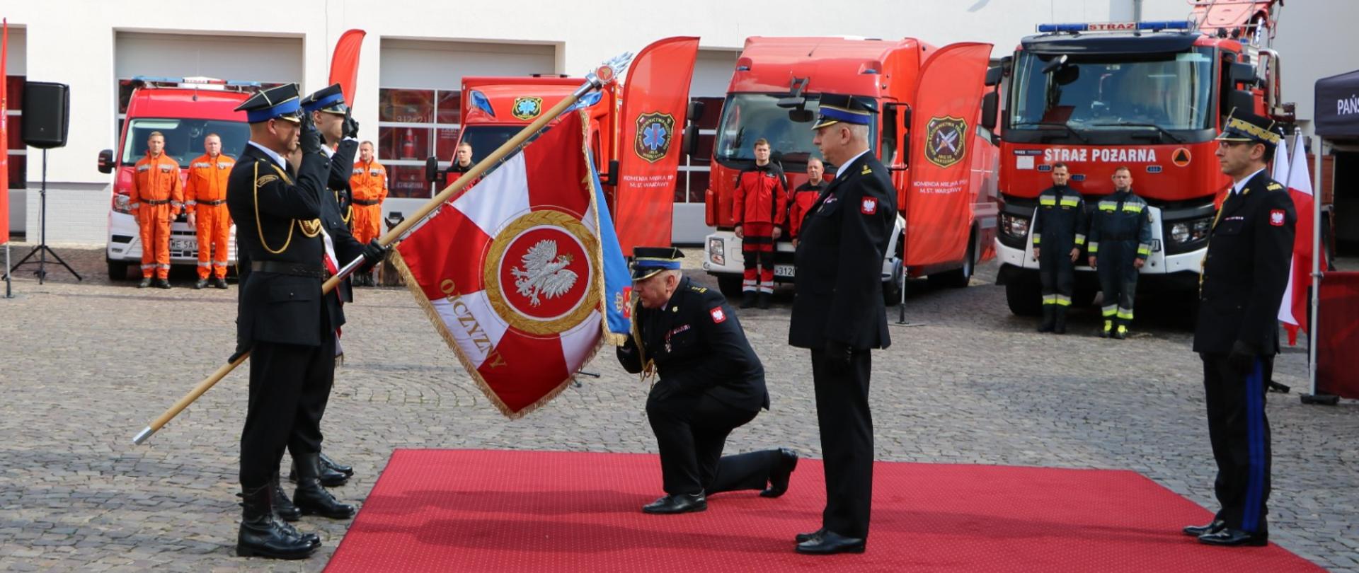 Na placu stoi kilku strażaków w mundurach galowych. Jeden z nich klęczy na czerwonym dywanie przed sztandarem, któremu oddaje honor. W tle widoczne czerwone samochody strażackie i biały budynek, a także stojacy na baczność strażacy.