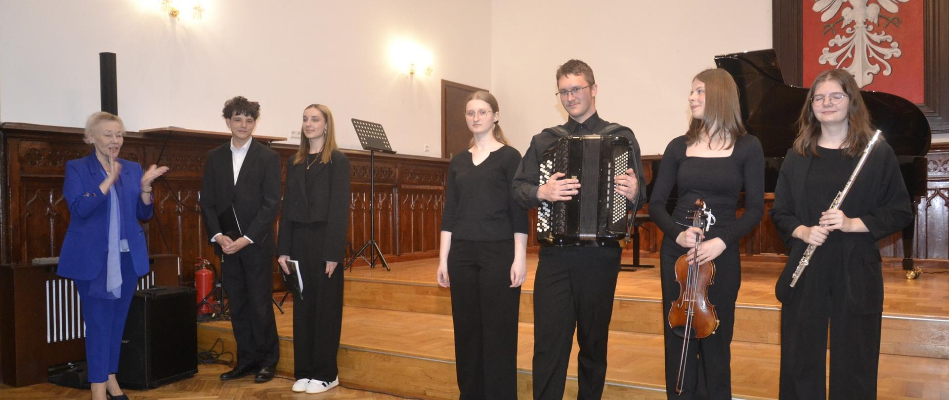 Uczniowie stojący na scenie z instrumentami podczas koncertu klasowego w Sali Królewskiej, prowadzonego przez dwoje uczniów.