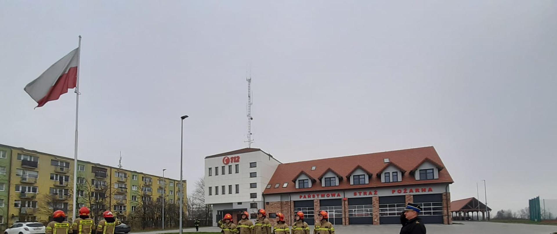 Na placu Komendy Powiatowej Państwowej Straży Pożarnej w Świeciu przed masztem na który została wciągnięta flaga narodowa stoi poczet flagowy, po prawej widać stojących w szeregu strażaków, a na przeciwko masztu stoi Dowódca Jednostki Ratowniczo-Gaśniczej bryg. Paweł Puchowski i oddaje honory. 