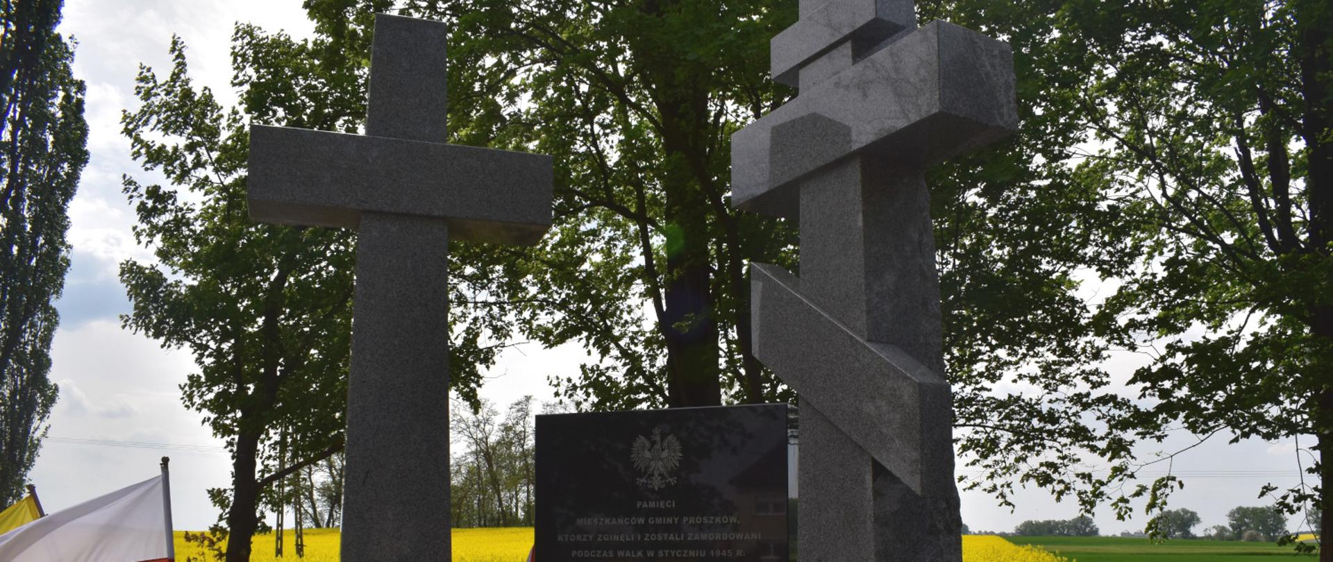 Na zdjęciu Pomnik w Folwarku - dwa krzyże a w ich środku tablica pamiątkowa. W tle widoczna łąka, po lewej stronie kadru flaga RP.
