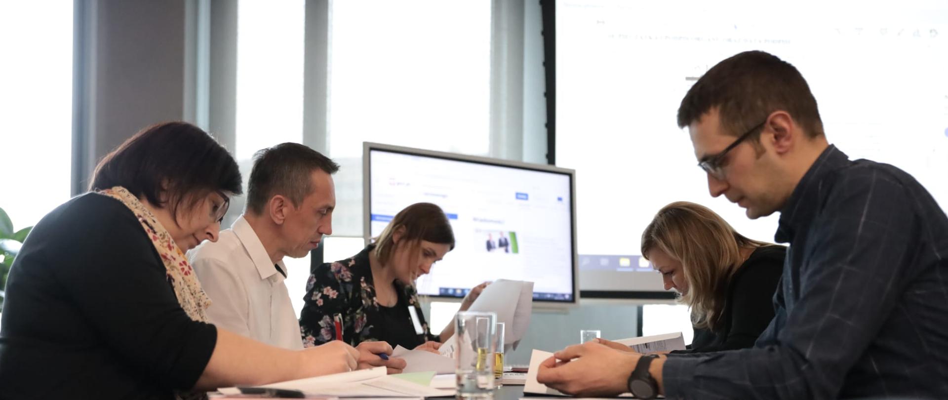 Warsztaty Ministerstwa Rozwoju i Technologii dla organów administracji architektoniczno-budowlanej - uczestnicy w trakcie warsztatów - siedzą przy stole pisząc i przyglądając się dokumentom - w tle ekran z prezentacją