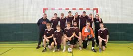 Grupa zawodników w sportowych strojach pozuje do zdjęcia na tle bramki, przed nimi leży piłka nożna.