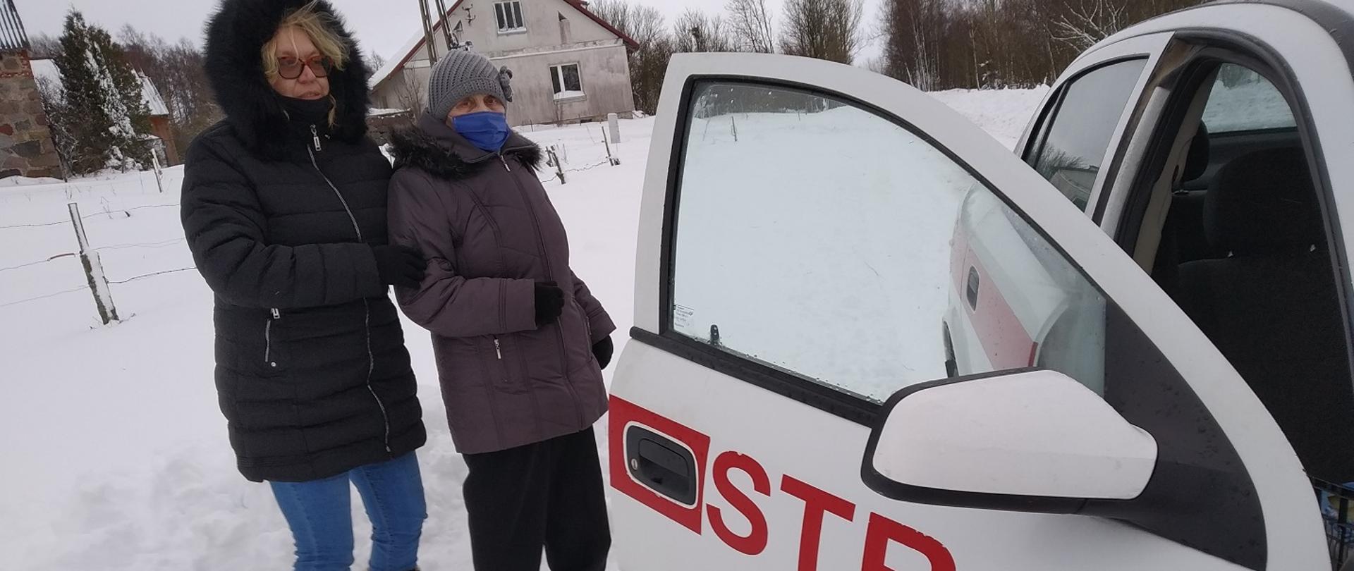 Po prawej stronie zdjęcia widać część samochodu strażackiego z otwartymi przednimi drzwiami od pasażera, samochód w kolorze białym z czerwonym pasem i napisem straż na otwartych drzwiach. Obok samochodu stoją dwie kobiety w zimowym ubraniu, na ziemi leży śnieg, w oddali widać słup energetyczny i budynek mieszkalny ustawiony szczytem do samochodu. 