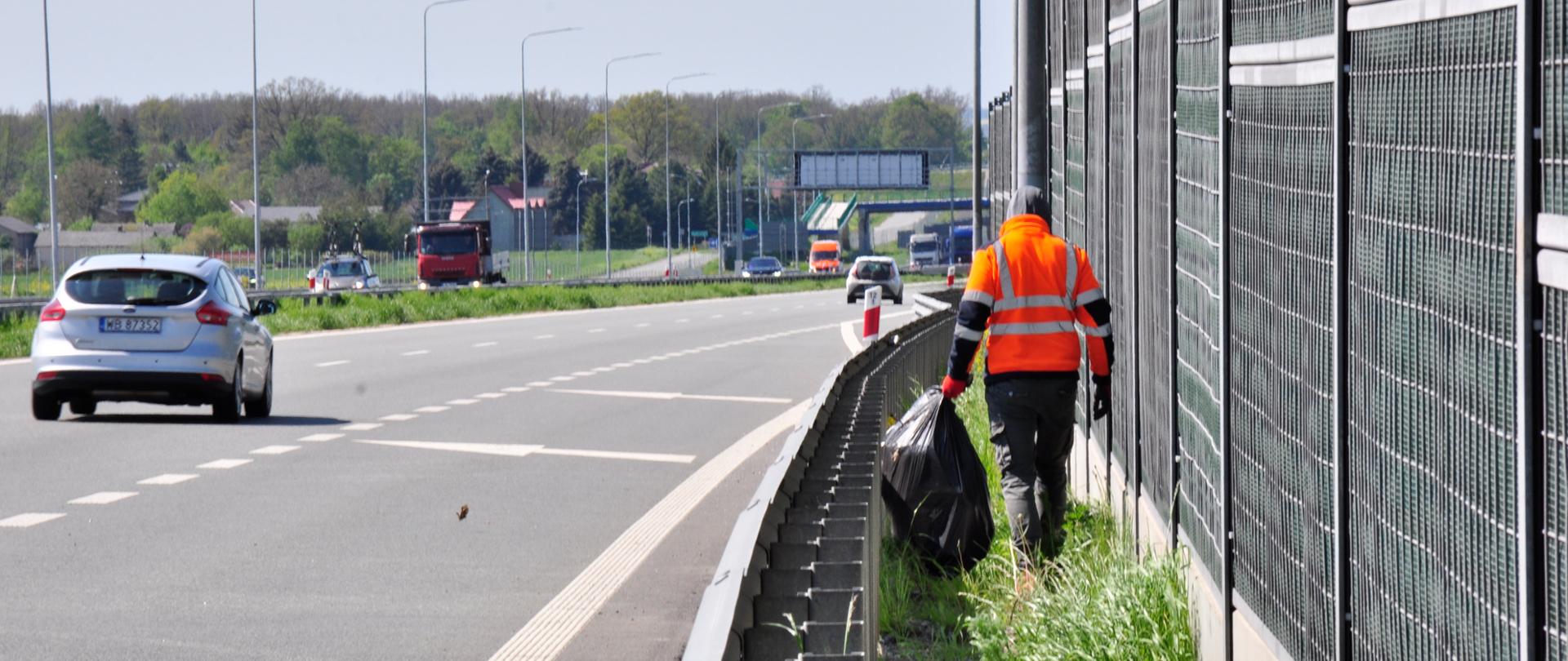 Zdjęcie przedstawia osobę ubraną w pomarańczową kamizelkę odblaskową, która porusza się wzdłuż pobocza autostrady, zbierając śmieci. Osoba ta trzyma duży czarny worek na śmieci i idzie obok metalowego ogrodzenia dźwiękochłonnego. Na drodze w tle widoczne są pojazdy, w tym srebrny samochód osobowy oraz czerwona ciężarówka. Po lewej stronie zdjęcia znajduje się pas zieleni oddzielający jezdnię od pobocza. W tle widoczne są drzewa oraz budynki, a dzień jest pogodny, z lekkim zachmurzeniem.