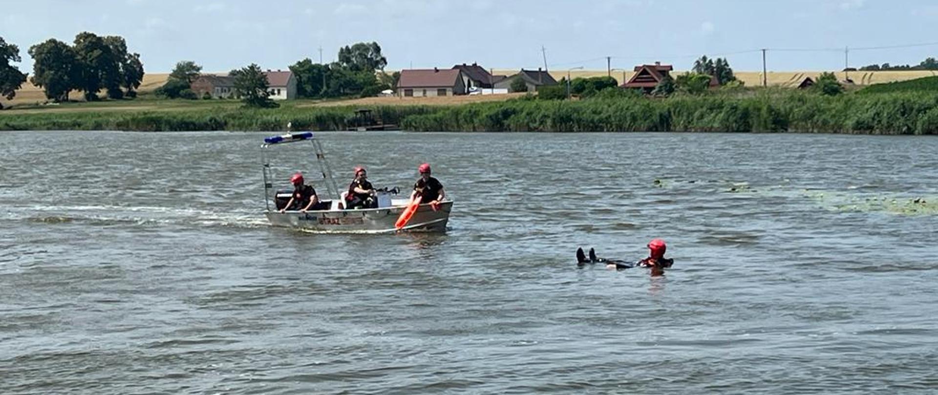 Na zdjęciu trzech strażaków na łodzi strażackiej na akwenie podpływająca do pozoranta w wodzie
