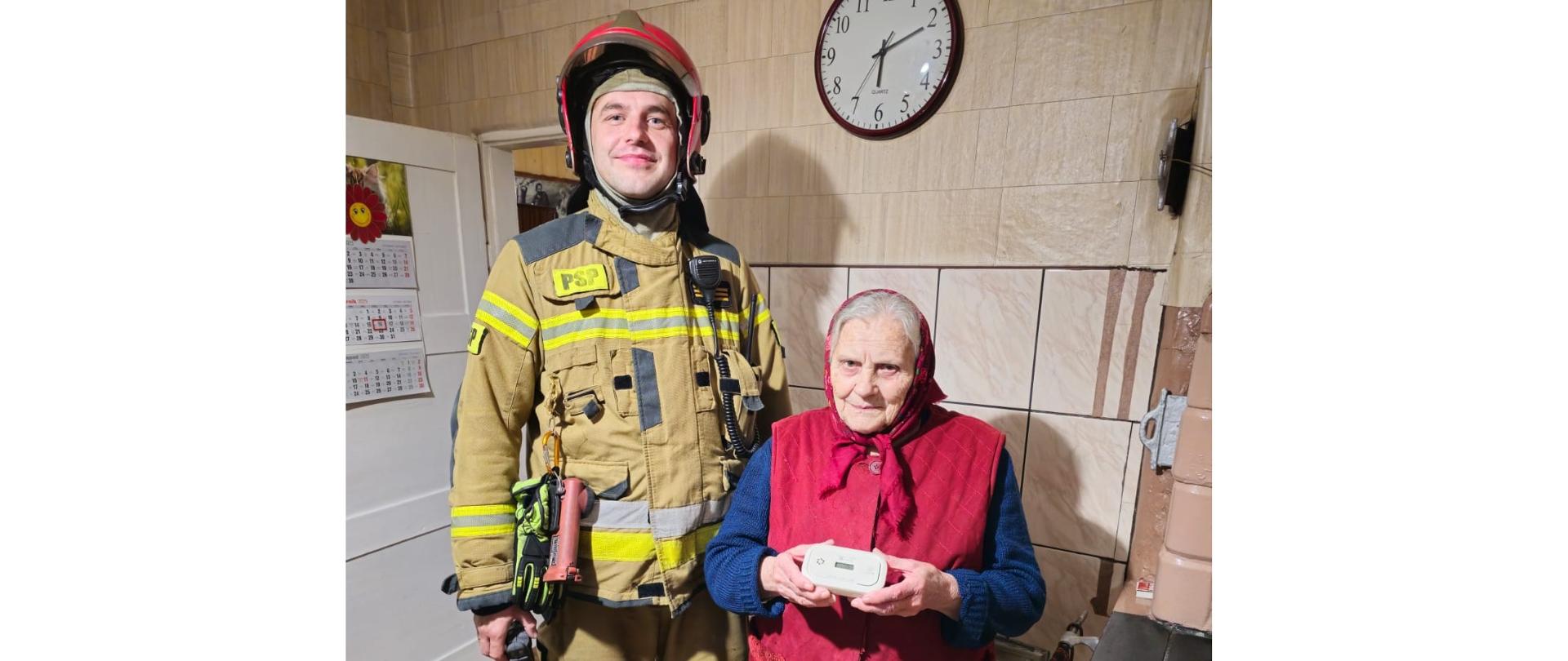 Zdjęcie przedstawia strażaka Komendy Powiatowej Państwowej Straży Pożarnej w Gołdapi, stojący obok mieszkanki lokalu, w którym uruchomiła się czujka tlenku węgla alarmując o zagrożeniu. Kobieta trzyma oburącz czujkę tlenku węgla