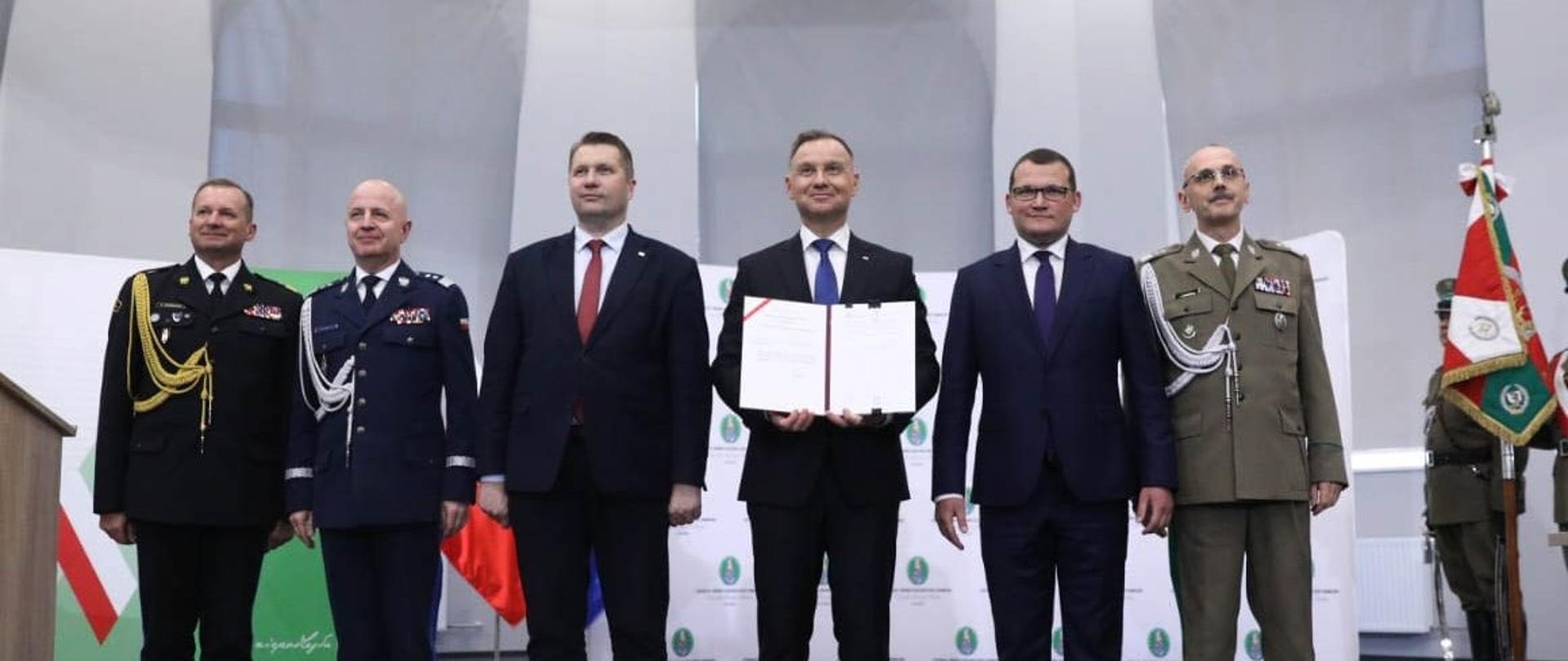 Sześciu mężczyzn stoi na środku sceny. Dwóch w granatowych mundurach, trzech w garniturach i jeden w zielonym mundurze. W środku mężczyzna w garniturze trzyma podpisany dokument.