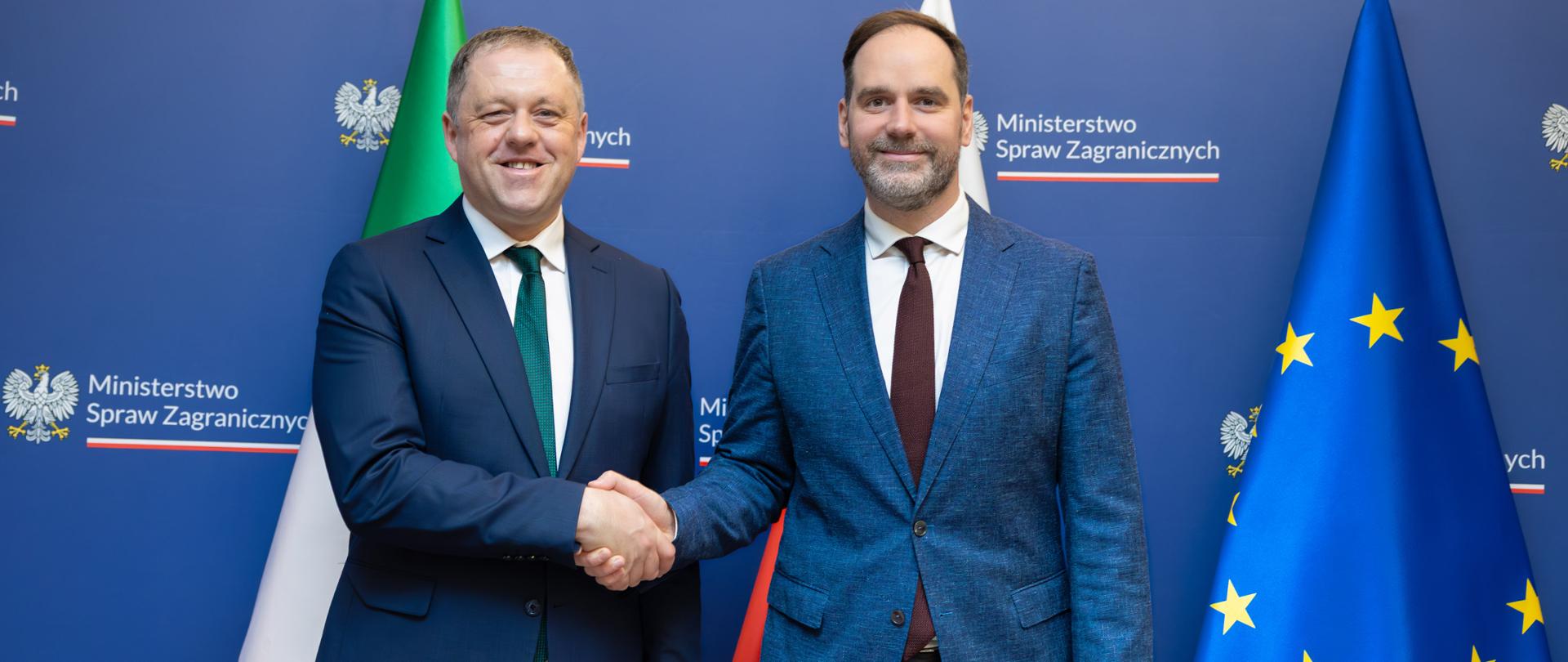 The photo shows two men in suits. They are standing, shaking hands, and facing the camera. Behind them are three flags: those of Ireland, Poland, and the European Union. 