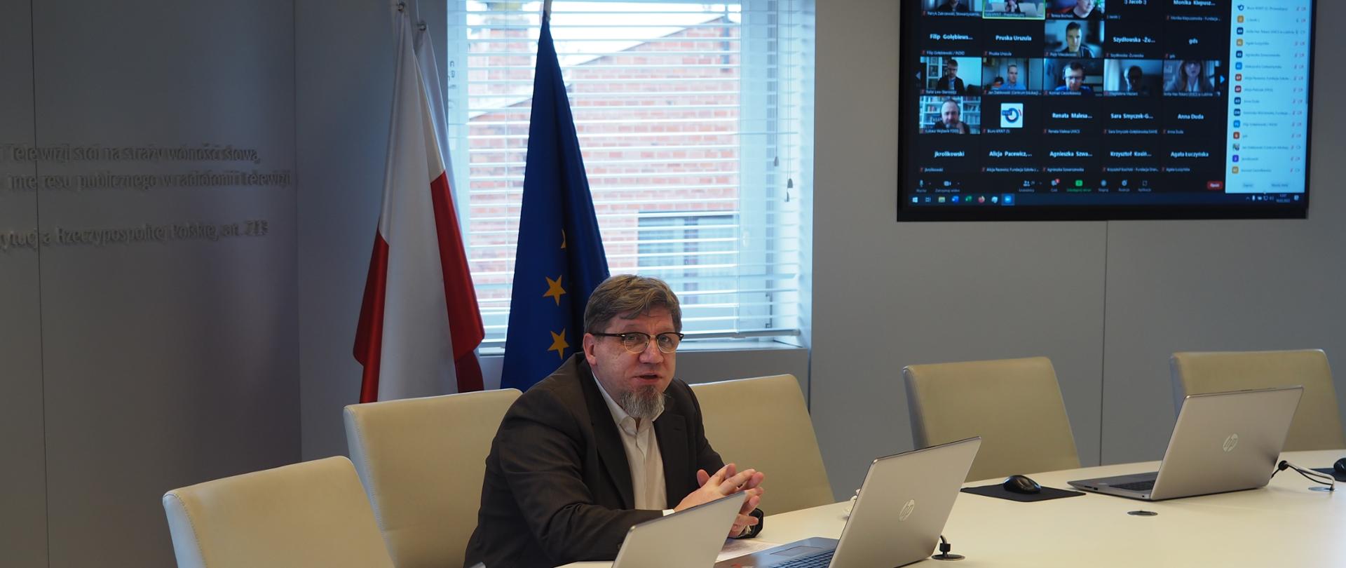 Zdjęcie ilustracyjne w poziomie. Przewodniczący KRRiT siedzi za stołem. W tle flaga Polski i wiszący na ścianie ekran. 