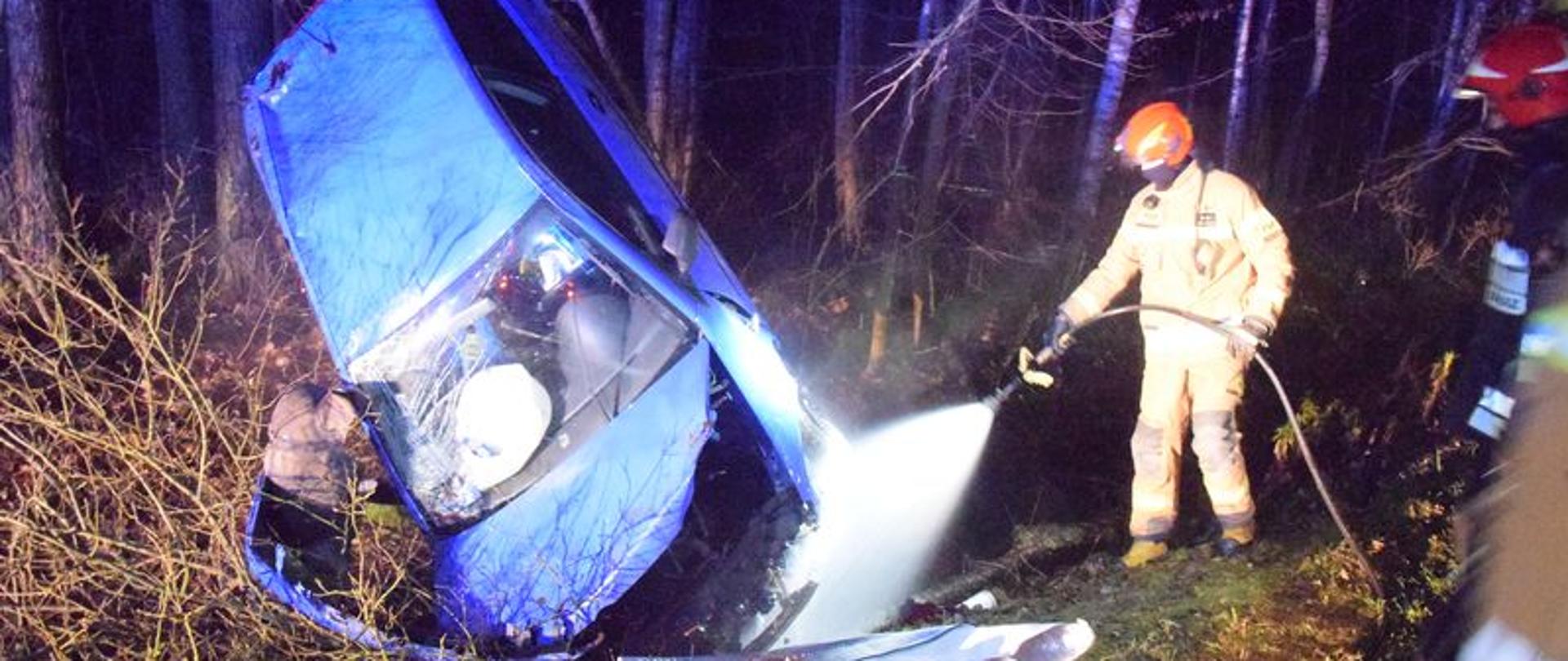 Na zdjęciu po lewej rozbity niebieski samochód osobowy, po prawej strażak ubrany w ubranie specjalne podaje prąd wody w kierunku rozbitej komory silnika.