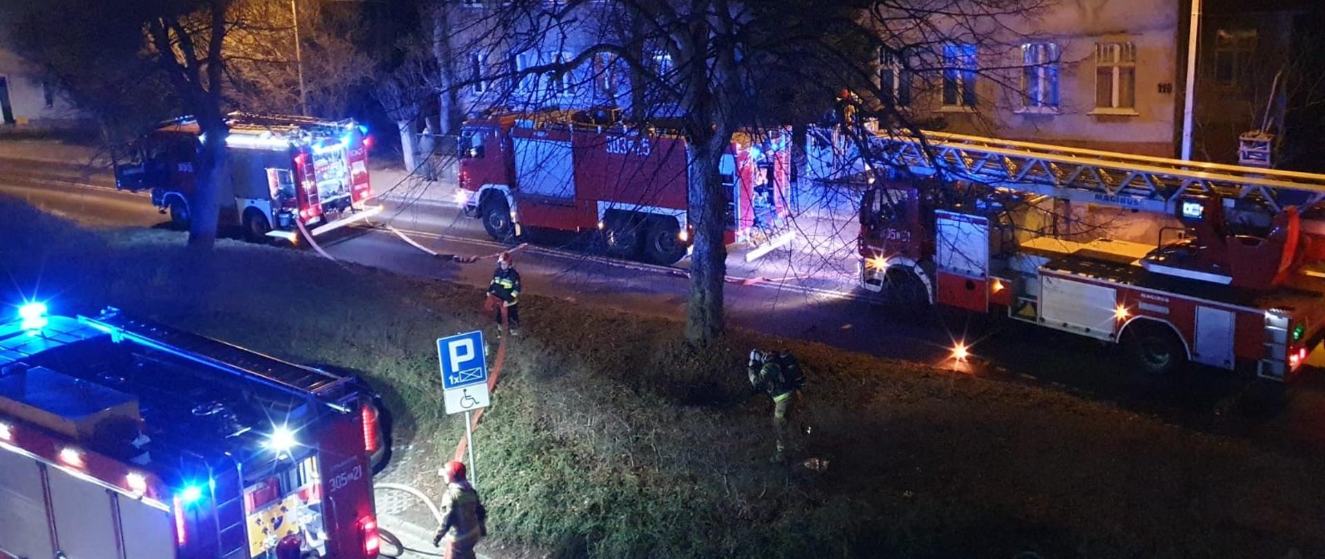 Pożar mieszkania przy ul. Rostockiej, wiać strażaków rozwijających węzę pożarnicze oraz cztery samochody pożarnicze stojące przy budynku.