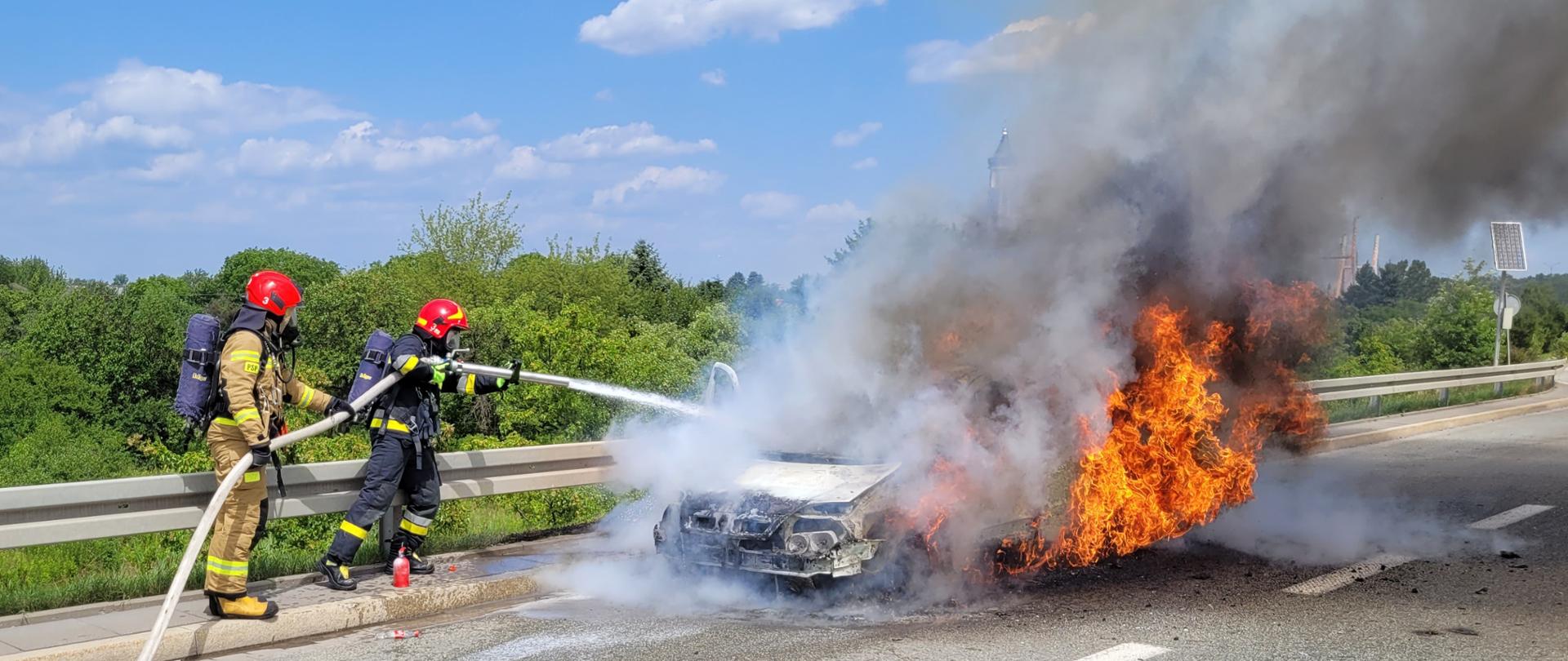  Na zdjęciu widać palący się samochód osobowy i gaszących go dwóch strażaków.
