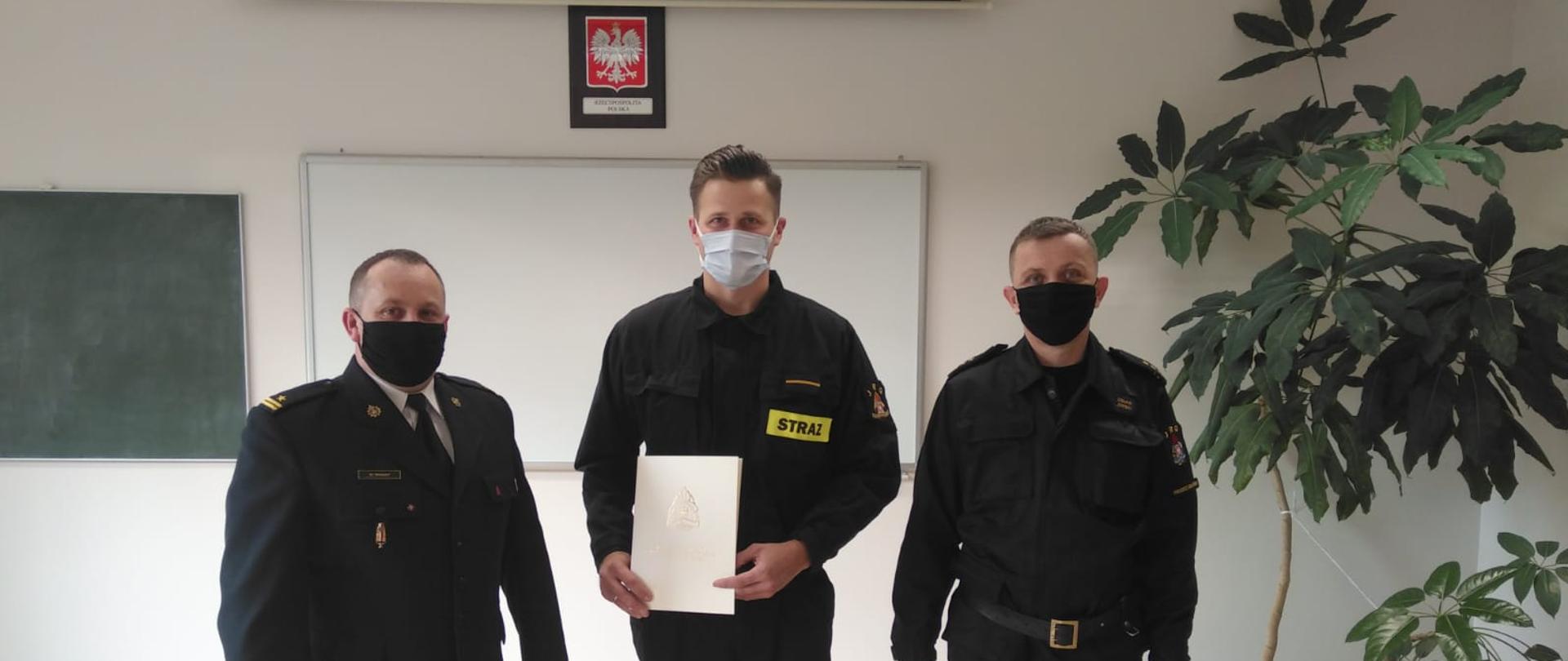 Obraz przedstawia pomieszczenie świetlicy na terenie Komendy Powiatowej. Na pierwszym planie z –ca Komendanta Powiatowego , dowódca JRG, awansowany strażak W uroczystości bierze udział zmiana służbowa. 