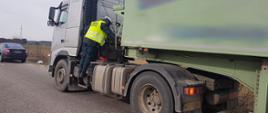 Na pierwszym planie: umundurowany inspektor podlaskiej Inspekcji Transportu Drogowego stoi na ciągniku siodłowym i sprawdza, czy do impulsatora skrzyni biegów (czujniku ruchu) jest przyłożony magnes neodymowy. W tle: przed zatrzymanym do kontroli zestawem ciężarowym stoi nieoznakowany radiowóz podlaskiej Inspekcji Transportu Drogowego z włączonymi niebieskimi światłami uprzywilejowania w ruchu drogowym.