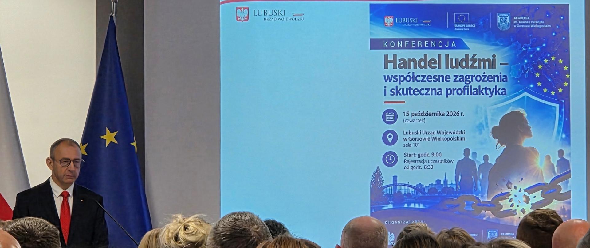 po lewej stronie stoi mężczyzna w garniturze, obok wyświetlona prezentacja z tytułem: "Handel ludźmi - współczesne zagrożenia i skuteczna profilaktyka