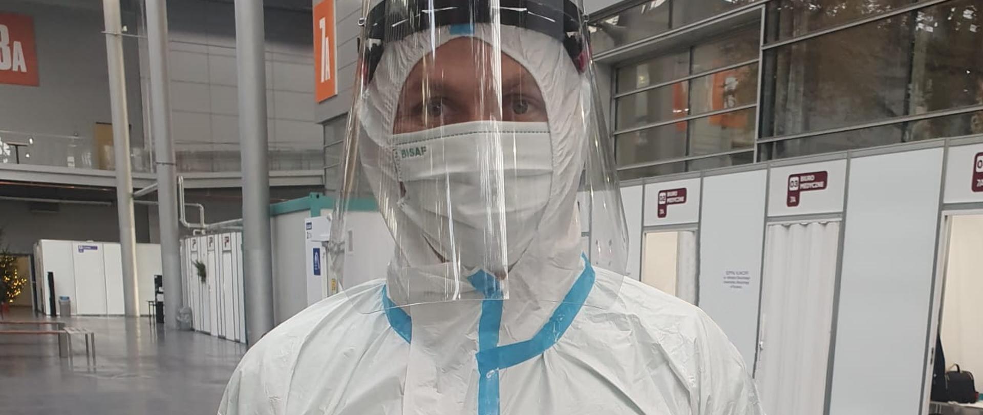 Ratownik ubrany w kombinezon ochronny, w tle boksy szpitala tymczasowego.