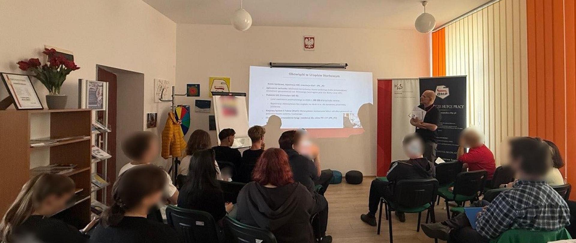 Uczestnicy siedzą w sali. Przed nimi stoi ekspert. Na ekranie wyświetlana jest prezentacja.
