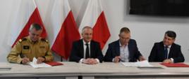 Strażak wojewoda pomorski oraz przedstawiciele firmy Enspirion siedzą przy stole i podpisują umowę za nimi znajdują się flagi Polski na ścianie wisi telewizor.