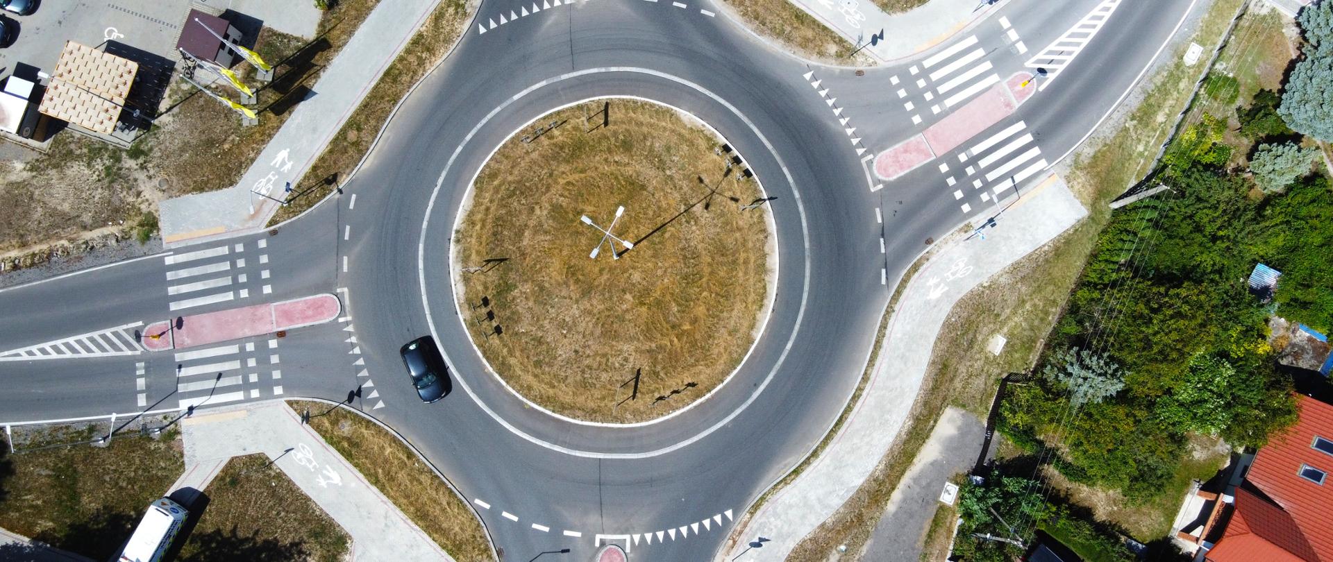 Widok z lotu ptaka na jednopasmowe rondo z wyspą centralną porośniętą trawą. Do ronda dochodzą cztery wloty z przejściami dla pieszych, drogami dla rowerów i chodnikami. Na jezdni widoczny jest jeden samochód, a wokół ronda znajdują się zabudowania i tereny zielone.