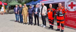 Grupa przedstawicieli służb ratowniczych, mundurowych i instytucji publicznych stoi przed namiotami wystawienniczymi podczas wydarzenia plenerowego. Wśród uczestników znajdują się ratownicy medyczni w pomarańczowych uniformach, funkcjonariusz policji, strażak oraz osoby w stroju cywilnym. W tle widoczne są stoiska informacyjne różnych organizacji, w tym Polskiego Czerwonego Krzyża, a także drzewa i teren rekreacyjny. Zdjęcie przedstawia oficjalne spotkanie i prezentację służb zaangażowanych w działania na rzecz bezpieczeństwa i ochrony zdrowia mieszkańców.