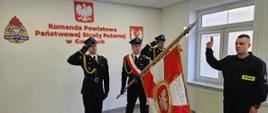 Na zdjęciu w mundurach galowych po lewej stronie poczet sztandarowy ze sztandarem. Po prawej strażak składający ślubowanie w ubraniu czarnym koszarowym. Uroczystość ślubowania mam miejsce w sali konferencyjnej.
