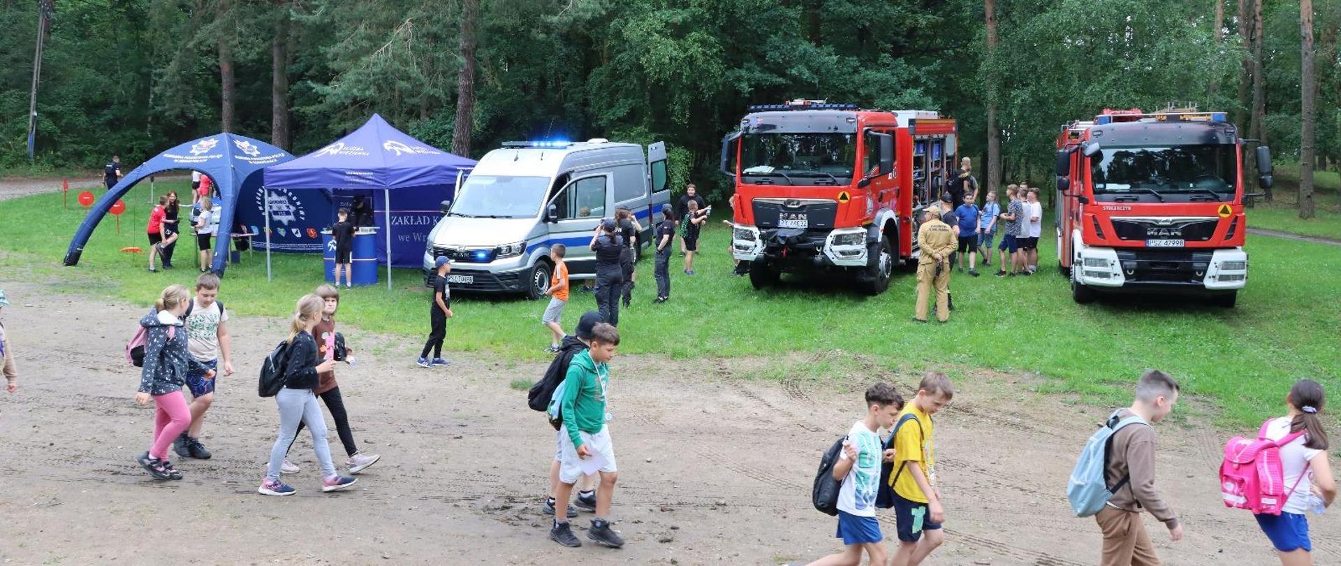 Na łące samochody strażackie i policja, dzieci maszerują