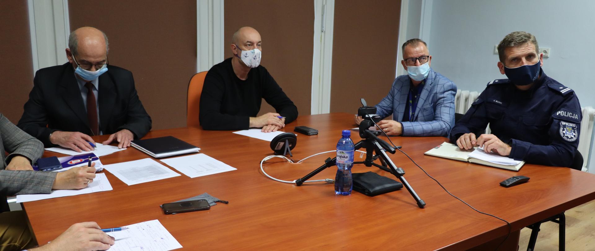 Ilustracja. W centralnej części kadru wojewoda opolski Adrian Czubak, podczas wideokonferencji ze starostami. Wojewoda spogląda w monitor, na twarzy ma ubraną maseczkę ochronną. Po jego prawej stronie zasiada dyrektor Wydziału Bezpieczeństwa i Zarządzania Kryzysowego, po prawej wicekurator oświaty. Po prawej stronie kadru widać policjanta. W lewej dolnej części kadru widać dłonie pozostałych uczestników spotkania.Wszyscy zasiadają przy dużym, prostokątnym stole.