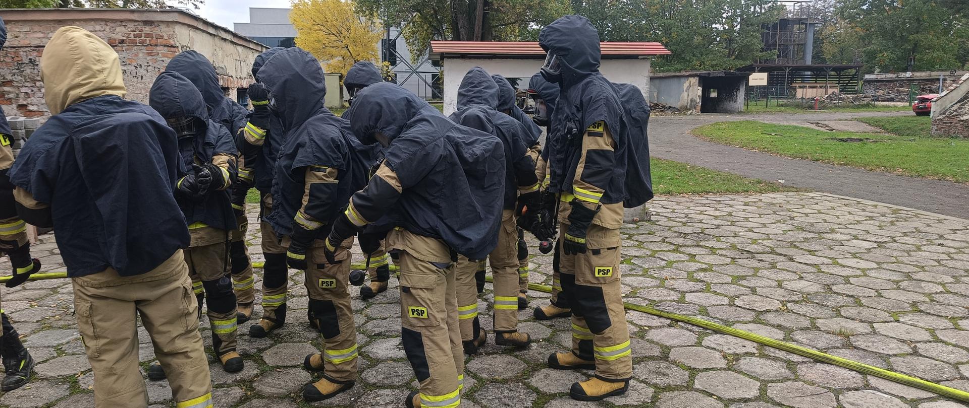 Poligon Centralnej Szkoły PSP, grupa kadetów z XXVIII turnusu dziennego studium aspirantów w umundurowaniu bojowym wraz z butlą oraz założonym ponczem ochronnym przed podejściem do ćwiczenia