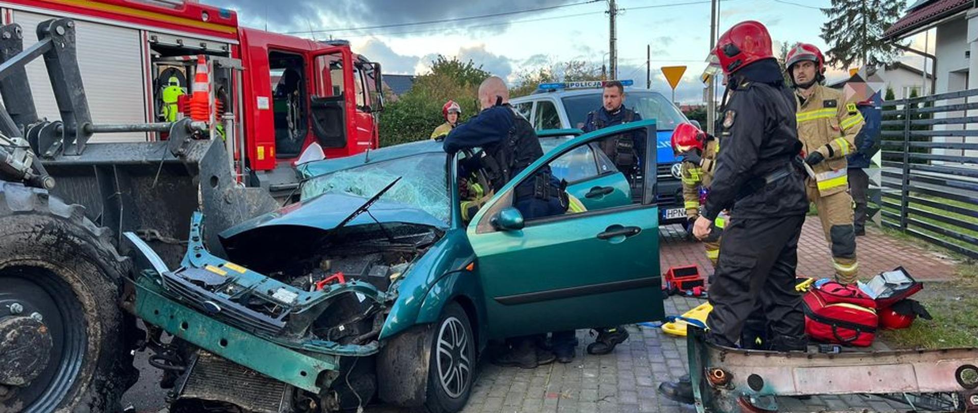 Zdjęcie przedstawia uszkodzony samochód osobowy, który zderzył się czołowo z koparko-ładowarką, strażaków prowadzący działania ratownicze oraz pomagający policjanci. W tle widoczny pojazd pożarniczy, samochód policji oraz sprzęt medyczny.
