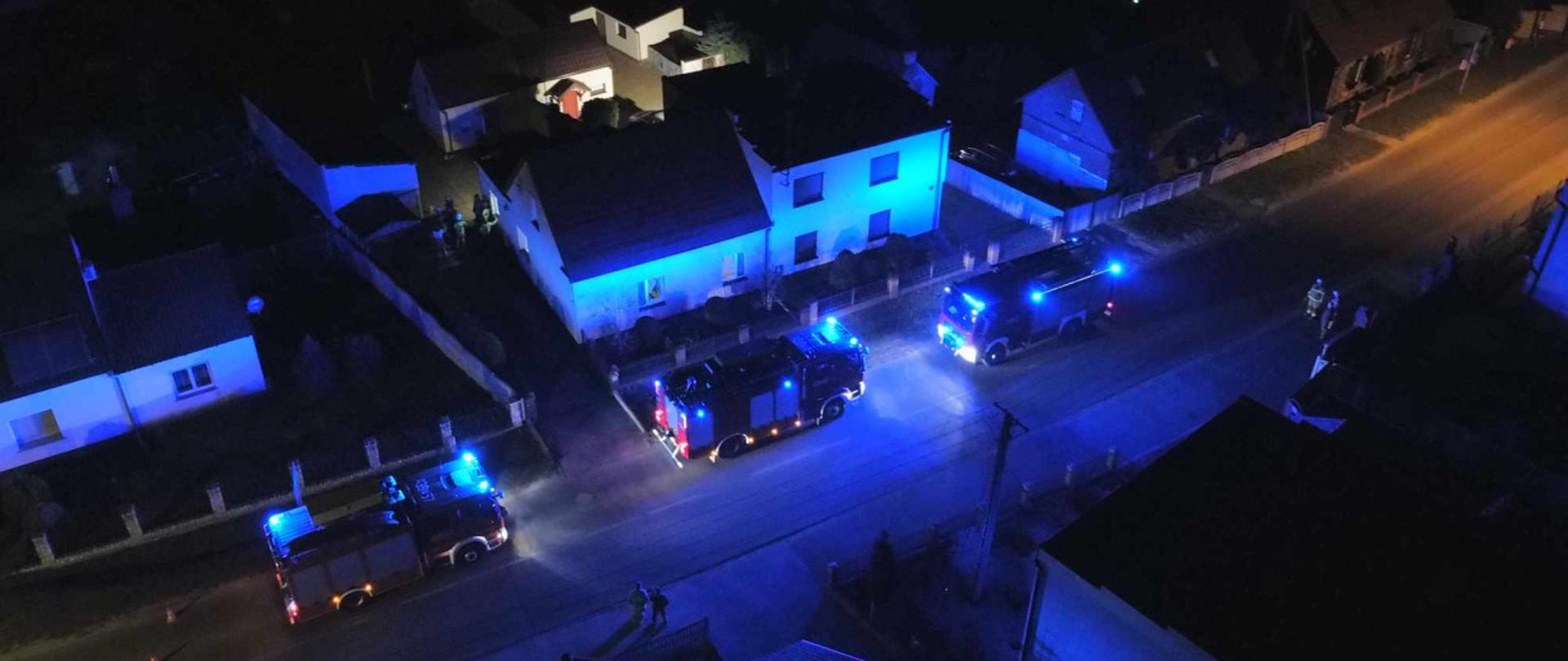 Trzy wozy strażackie z włączonymi światłami błyskowymi. Stoją one na drodze przy budynku.