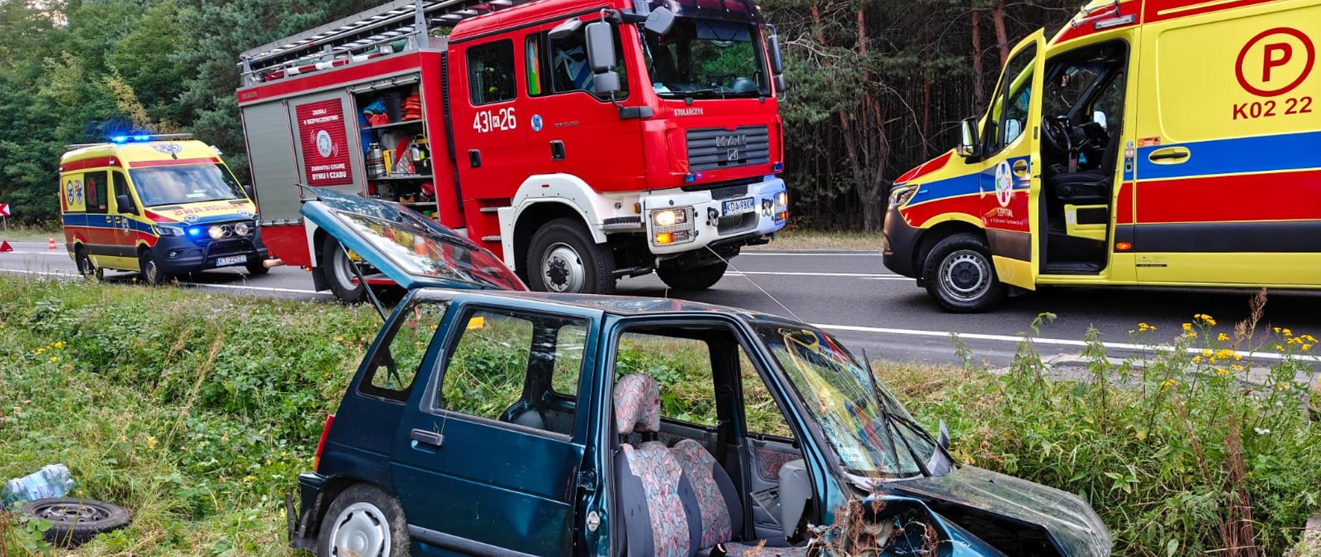 Na zdjeciu widać samochód osoby po wypadku, na drugim planie pojazdy ratownicze, uszkodzony przod pojazdu ktory uderzył w mostek przydrożny czołowo.