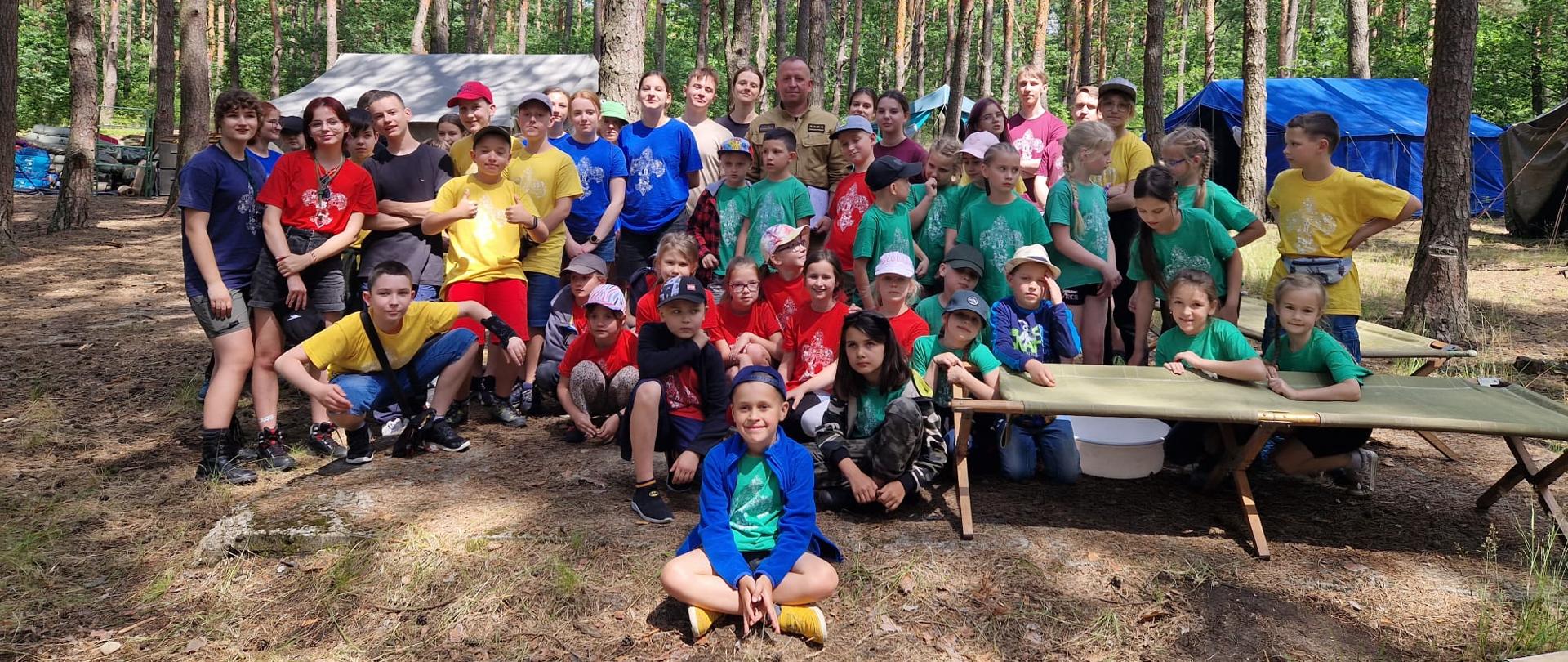 Zdjęcie grupowe uczestników obozu zrobione w leśnym obozowisku. w tle widać namioty oraz drzewa.