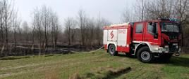 Zdjęcie przedstawiające pogorzelisko po pożarze trawy. Na pierwszym planie samochód straży pożarnej oraz rozwinięta linia gaśnicza.