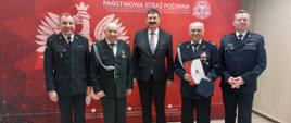 Zdjęcie przedstawia komendantów, ministra oraz druhów, którzy otrzymali świadczenia ratownicze. Osoby stoją przed banerem komendy. 