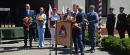 Plac przed komendą. Przy mównicy stoi wicestarosta rawicki, który przekazuje strażakom kolicznościowe życzenia. W tle, z bukietami kwiatów w rękach stoją samorządowcy.