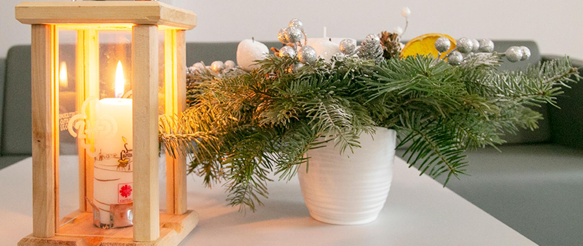 Na stole Światełko Betlejemskie 2020. Obok ozdobna kompozycja z iglastych gałązek i bombki. 