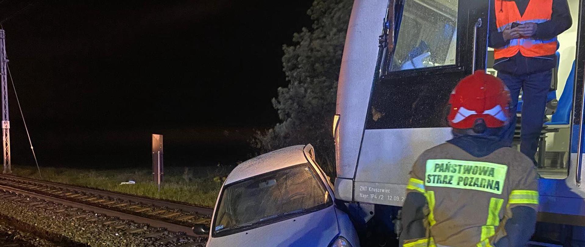 Na zdjęciu znajduje się wrak samochodu, pociąg, strażak w ubraniu specjalnym oraz kolejarz