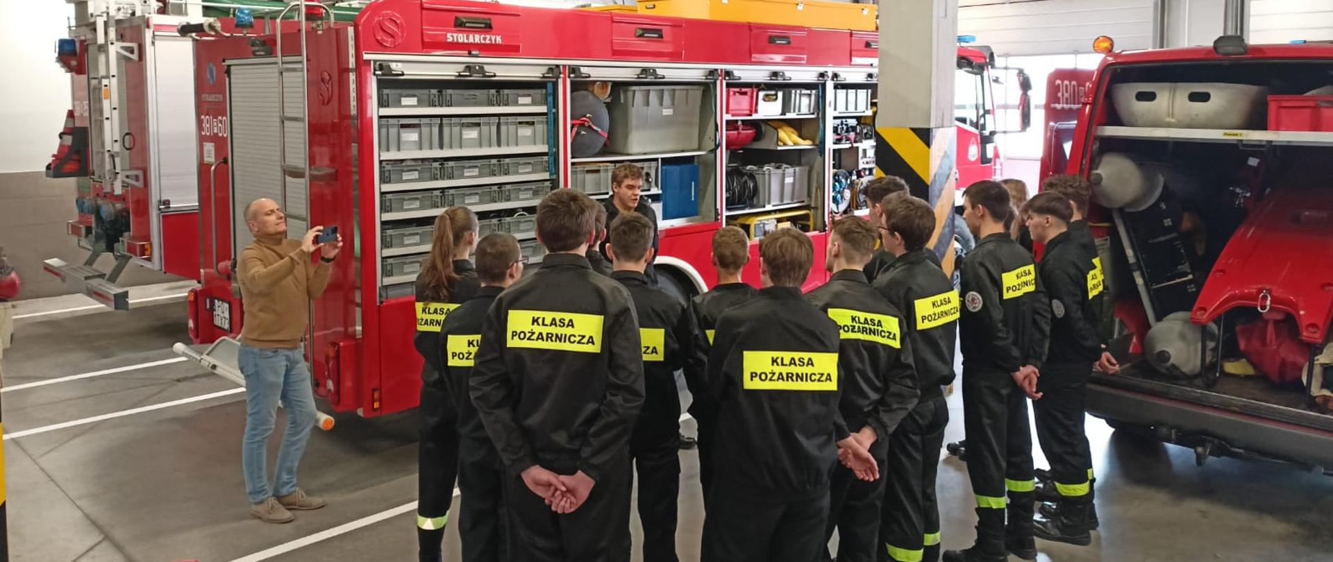 Strażacy i młodzież w mundurach stoją na garażu miedzy samochodami pożarniczymi.