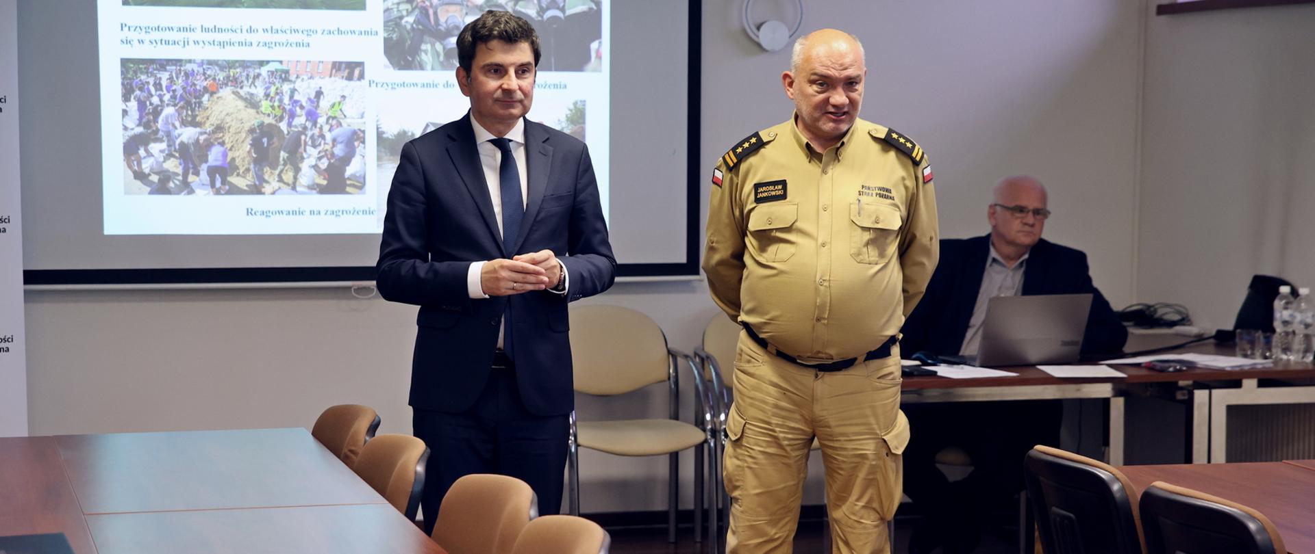 Sala konferencyjna podczas rozpoczęcia szkolenia z zakresu ochrony ludności i obrony cywilnej, od lewej stoją wojewoda śląski oraz komendant Centralnej Szkoły PSP, w tle widoczny ekran multimedialny z wyświetlonym slajdem prezentacji. W rogu po prawej siedzący przy biurku prelegent