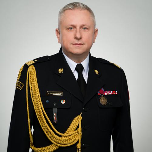 Portret mężczyzny w galowym mundurze Państwowej Straży Pożarnej, sfotografowanego od pasa w górę na jasnym, jednolitym tle. Ma krótkie, siwe włosy i patrzy bezpośrednio w obiektyw. Ubrany jest w czarny mundur galowy z białą koszulą i czarnym krawatem. Na mundurze widoczne są dystynkcje, złote emblematy, odznaczenia i baretki, a także tabliczka z nazwiskiem „S. Sierpatowski”. Przez prawe ramię przewieszony jest ozdobny złoty sznur galowy. Postawa jest wyprostowana i oficjalna, a całość ma charakter reprezentacyjnego portretu służbowego.