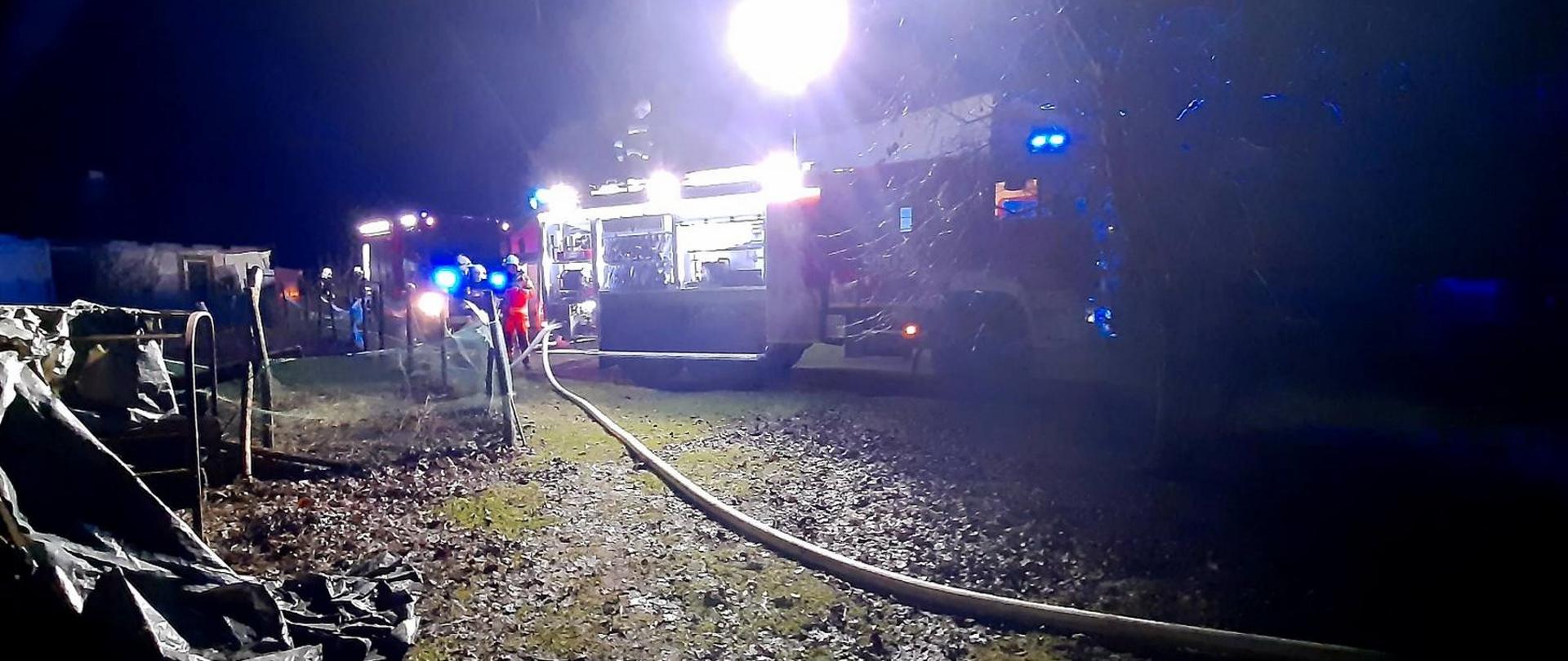 Obok ogrodzenia posesji ustawione są pojazdy pożarnicze od których podawana jest woda wężami pożarniczymi do pożaru palącej się wiaty. 