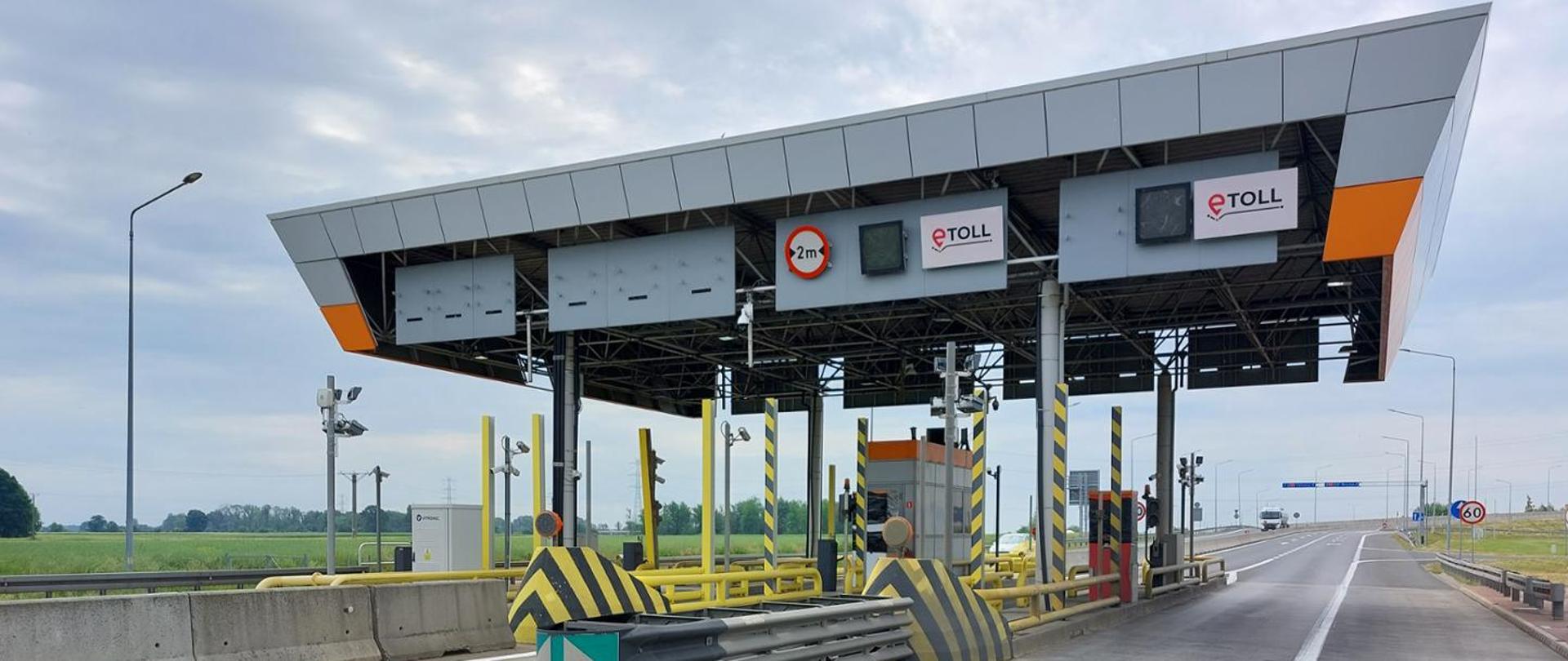 Plac poboru opłat na autostradzie A4 widziany z poziomu jezdni. Zadaszona konstrukcja z oznaczeniami systemu e‑TOLL obejmuje kilka pasów ruchu z barierkami i urządzeniami kontrolnymi. Po prawej stronie widać pustą drogę z ograniczeniem prędkości do 60 km/h, a w tle płaski krajobraz z polami i pochmurnym niebem.