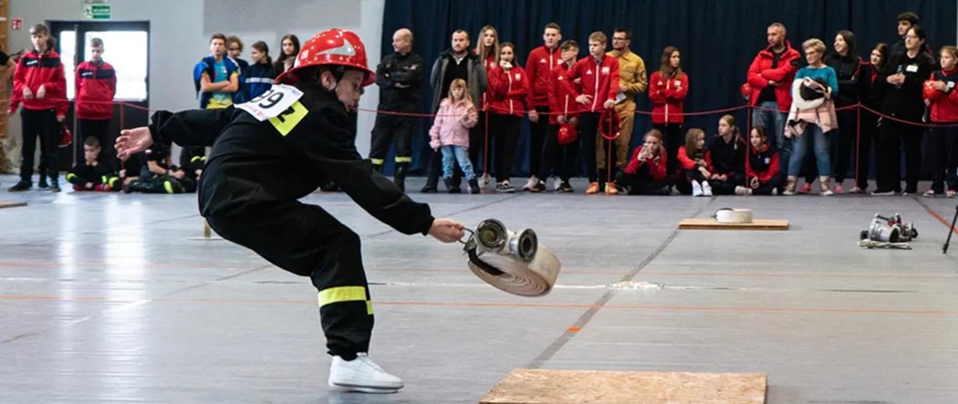 Widok z boku - zawodniczka w trakcie wykonywania konkurencji na hali sportowej, w tle kibice.