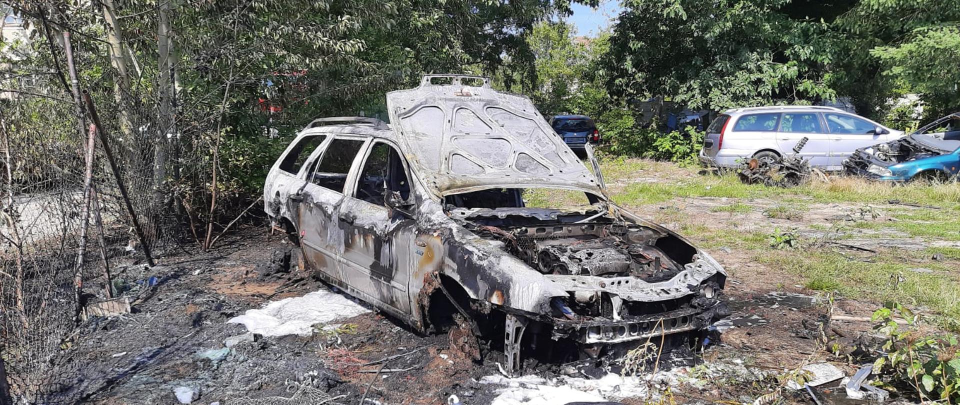 Zdjęcie przedstawia samochód osobowy z podniesioną maską, który objęty był pożarem. W oddali widoczne są samochody oraz części samochodowe.