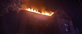 Na zdjęciu widać pożar dachu murowanego budynku mieszkalnego w nocy. Z dachu wydobywają się intensywne płomienie i gęsty dym, oświetlając okolicę pomarańczowym blaskiem. Budynek jest ciemny, a okna na niższych kondygnacjach wyglądają na puste lub zniszczone. Po lewej stronie widać gałęzie drzewa, a w tle słabe światła uliczne. Cała scena ukazuje poważne zagrożenie pożarowe.