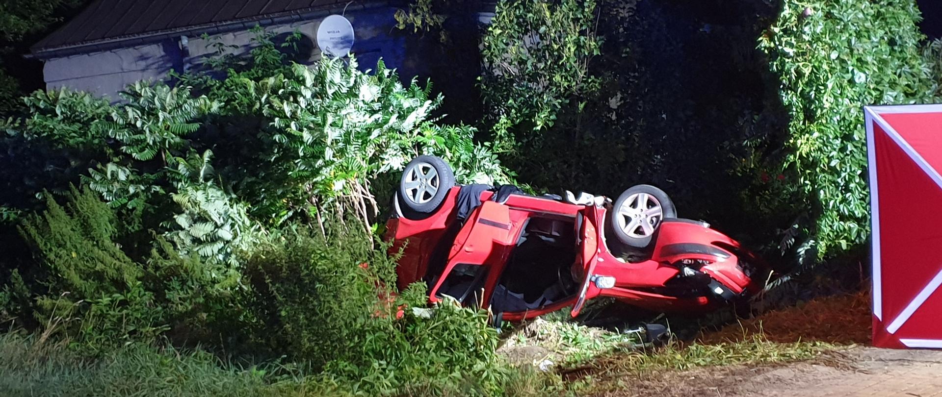 Na zdjęciu widać uszkodzony pojazd osobowy. Zdjęcie wykonane w nocy. Sztuczne oświetlenie. Widać fragment pola. Auto w kolorze czerwonym. Leży na dachu.