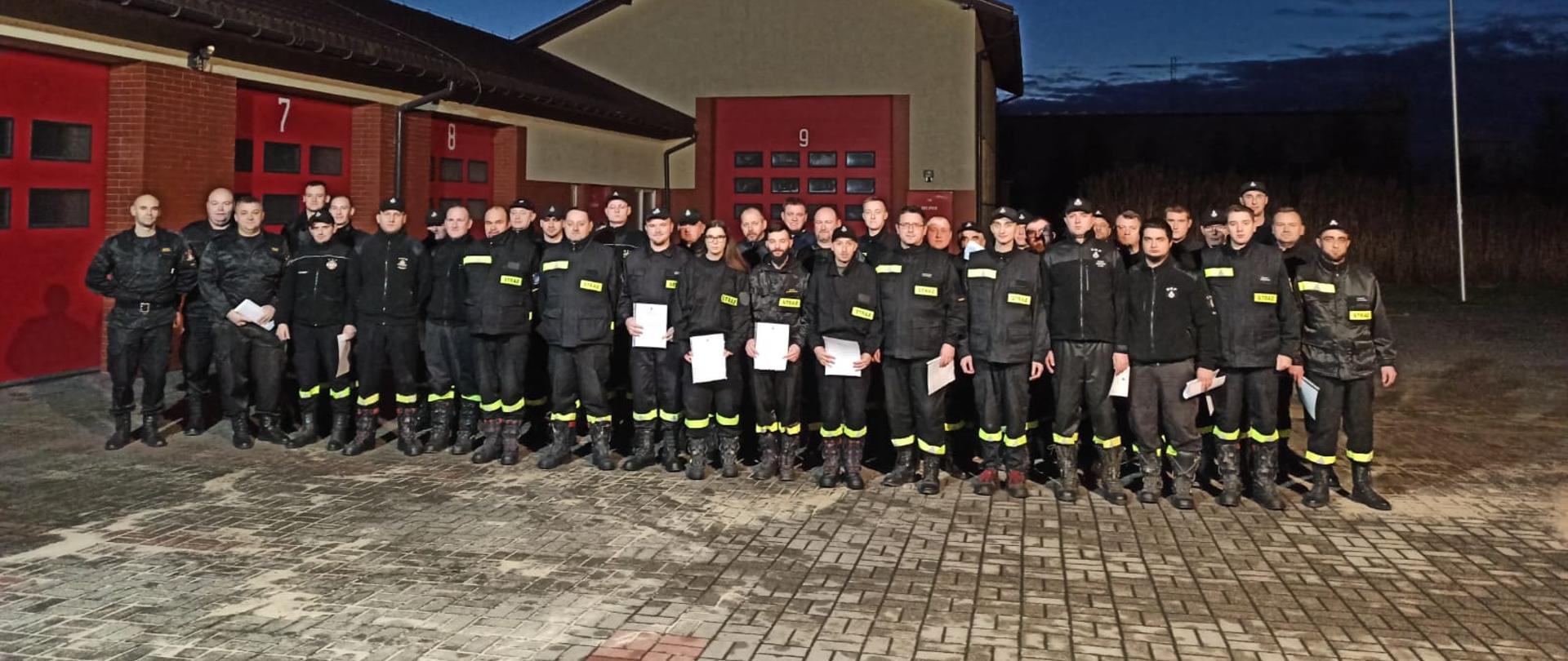 Strażacy stoją w dwuszeregu na placu z polbruku. W tle z tyłu i po prawej stronie budynki z czerwonymi bramami.