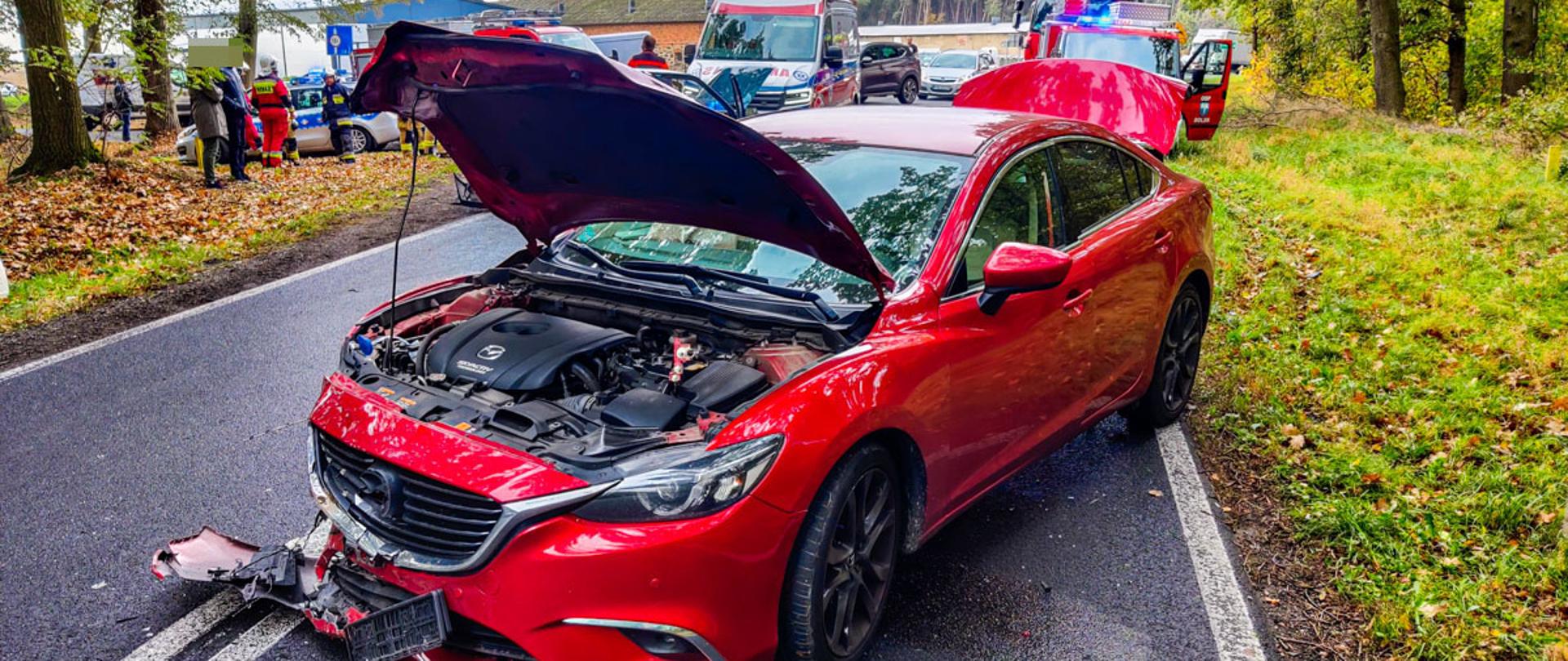 Na zdjęciu czerwony samochód osobowy rozbity przód z podniesioną maską za nim Policja ZRM i samochody ratowniczo - gaśnicze i strażacy